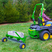 A person on a green riding lawnmower tows the John Deere 40 Spike Aerator with a steel weight tray filled with cinder blocks. Set against evergreen trees, they wear a green cap and red plaid shirt, navigating the grassy lawn with the power of galvanized steel stars.