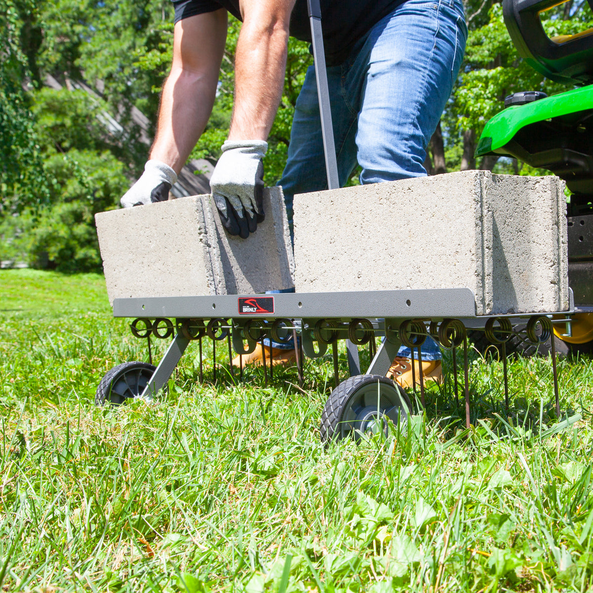 Outdoors, a person in gloves loads concrete blocks onto a Brinly Parts 40 Dethatcher in Hammered Gunmetal (DT2-40BH-S), which is on grass near a green lawnmower. The dethatcher resembles a wheeled lawn aerator with tines.