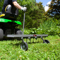 Wearing gloves, a person uses the Brinly Parts 40″ Dethatcher in Hammered Gunmetal (DT2-40BH-S) with metal tines and rubber wheels, to tow behind a lawn tractor across grassy terrain amidst serene trees and greenery.