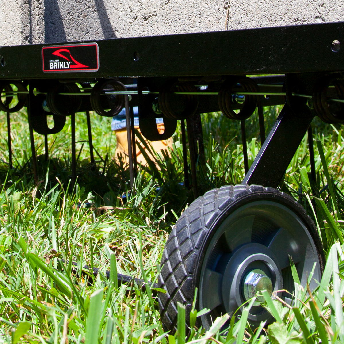 A Brinly Parts 40″ Tow-Behind Dethatcher (DT2-40BH-G) with metal tines and wheels is efficiently removing thatch as it’s pulled across a grassy lawn, with a persons shoe partially visible in the background.