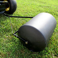 A Brinly Parts 28 Gallon Push or Tow Poly Lawn Roller | PRC-242BH, a black cylindrical heavy-duty polyethylene drum, is attached to a lawn mower with yellow-accented wheels, and rests on a grassy lawn with its metal frame connected to the mower.