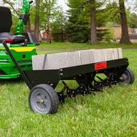 A garden tractor tows the Brinly Parts 48″ Tow-Behind Plug Aerator (PA-482BH) filled with concrete blocks over a grassy yard. The black aerator, with spiked wheels and steel tines, showcases the brand logo while enhancing the root zone. Trees and houses create a peaceful backdrop.