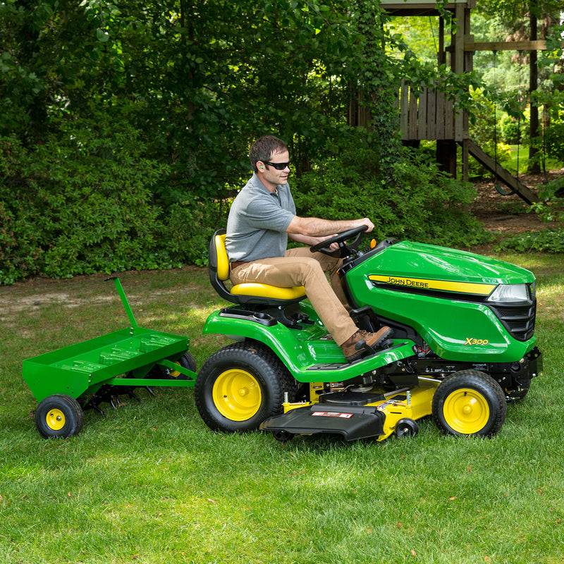 Wearing sunglasses, a person drives a green and yellow ride-on lawn mower with a green trailer, ideal for Brinly Parts Plug Aerator - 48 in Tow-Behind. They navigate a grassy lawn with trees and shrubs, effortlessly prepping the ground for aeration.