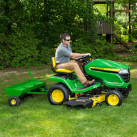 A person is mowing with a John Deere 48 Tow-Behind Plug Aerator equipped with carburized steel tines, wearing sunglasses, a gray shirt, and khaki pants on a sunny day amid lush trees.