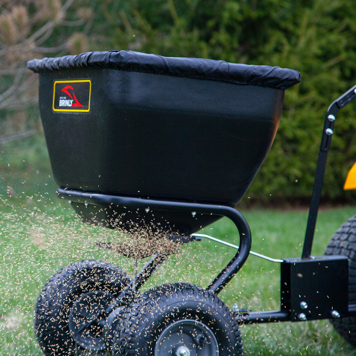 The Brinly Parts 175 LB. Tow-Behind Deluxe Spreader, model BS361BH-A, features four wheels, a rust-proof polyethylene container with the logo and a universal hitch for seamless integration while fertilizing or seeding your lawn against lush verdant green tree backdrops.