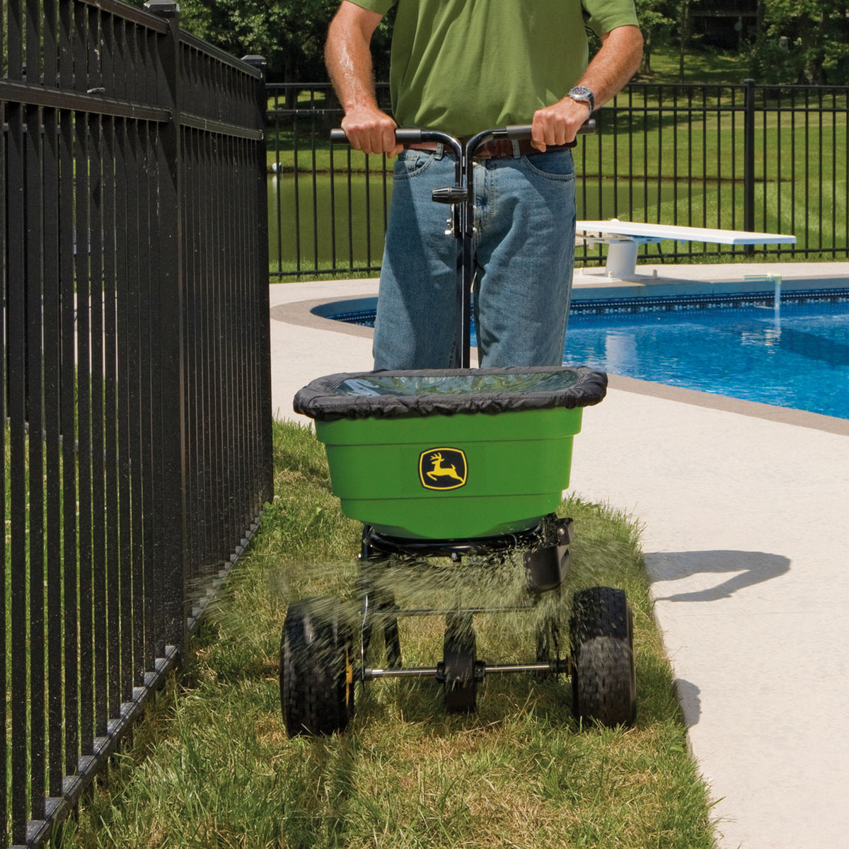 A person in jeans and a green shirt effortlessly maneuvers the John Deere All-Season Push Broadcast Spreader LP31340L-A by the poolside, scattering granules from its durable poly hopper with ergonomic split handles for easy control, as a black fence runs parallel to their path.