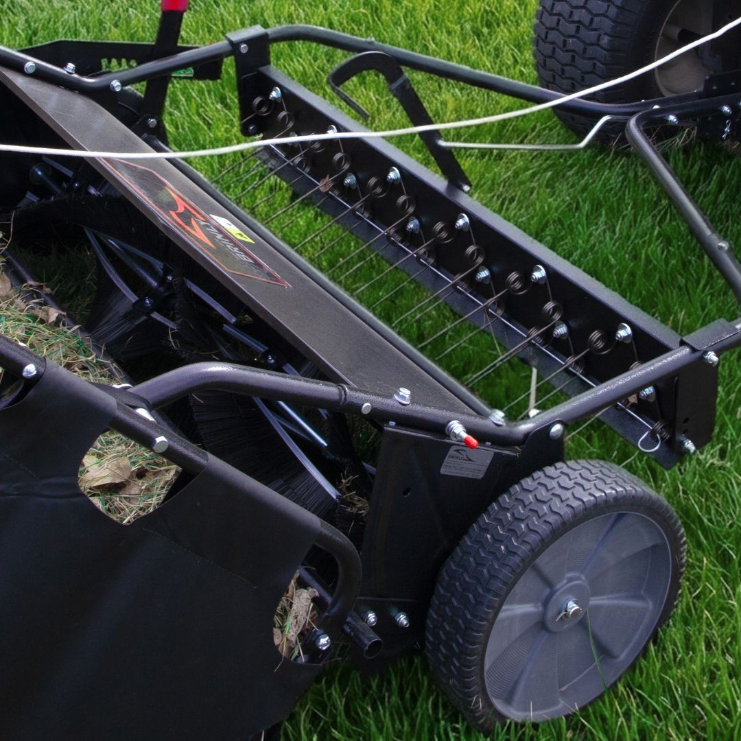 Close-up of the Brinly Parts 54″ Combo Lawn Sweeper with Dethatcher (LSDT2-54BH-G) on green grass, showing metal tines, wheels, and collection bag—a versatile 2-in-1 tool for effective yard maintenance.