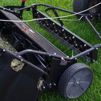 Close-up of the Brinly Parts 54″ Combo Lawn Sweeper with Dethatcher (LSDT2-54BH-G) on green grass, showing metal tines, wheels, and collection bag—a versatile 2-in-1 tool for effective yard maintenance.