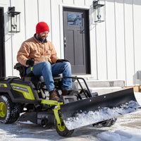 Wearing a red beanie and brown jacket, an individual expertly navigates a RYOBI Electric Zero-Turn Mower equipped with a Brinly Parts Front-Mount Blade (FB-42RYZT) to clear snow from the driveway. A white building with dark-framed windows and doors stands in the background.