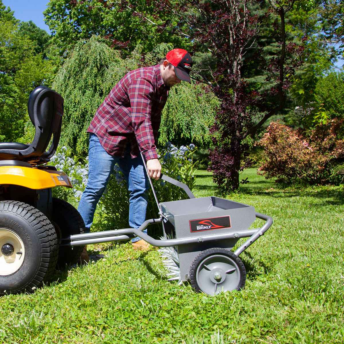 A person in a red cap and plaid shirt adjusts a Brinly Parts 40 Combination Aerator Spreader in Hammered Gunmetal, model AS2-40BH-S, attached to a yellow lawn tractor on a vibrant green lawn with trees and shrubs under a clear blue sky.