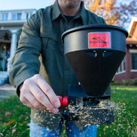 Wearing a green jacket, the person skillfully scatters fertilizer over their lawn using the Brinly Parts 5lb Capacity Handheld Spreader (HHS3-5BH), seeds dispersing easily.