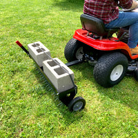 A person in jeans and a plaid shirt is using a red lawn tractor to tow a Brinly Parts 40 Tow-Behind Spike Aerator (SAT2-40BH-G) with two concrete blocks for extra weight, enhancing lawn aeration as it rolls across the grass.