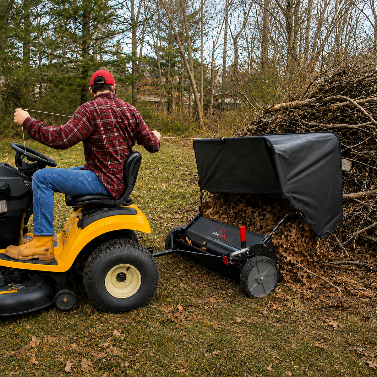 A person on a yellow lawn tractor uses the Brinly Parts 42″ Lawn Sweeper with DOUBLE-HELIX Brushes (LS2-42BH2-G) as a tow-behind attachment to efficiently and easily collect a large pile of brown leaves from a grassy, tree-filled yard.