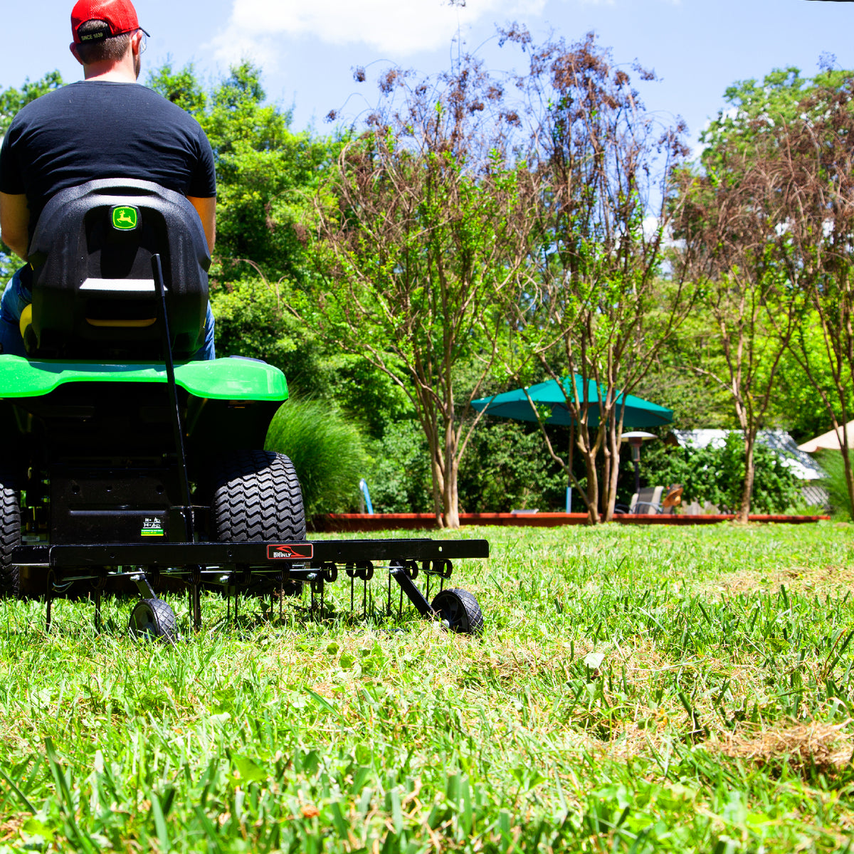 A person in a red cap skillfully uses a Brinly Parts 40″ Tow-Behind Dethatcher (DT2-40BH-G) on a green lawn mower across the grassy area, surrounded by trees. A small structure with a green roof is in the background under a sunny, partly cloudy blue sky.