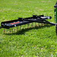 A Brinly Parts 48” Tow-Behind Dethatcher (DT-480BH) is pulled by a green lawn tractor across a grassy yard, with its metal tines visible as they remove thatch. A well-maintained green lawn is in the background.