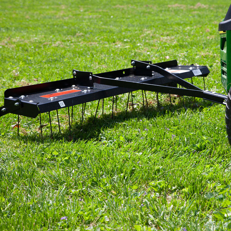 A Brinly Parts 48” Tow-Behind Dethatcher (DT-480BH) is pulled by a green lawn tractor across a grassy yard, with its metal tines visible as they remove thatch. A well-maintained green lawn is in the background.