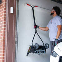 A man in safety gear hangs a Brinly Parts 20 Push Spike Aerator with 3D galvanized steel tines on a garage wall rack beside a brick wall and parked car, preparing for his next lawn aeration project.