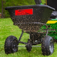 Attached to a green tractor, the Brinly Parts 125 Lb. Tow-Behind Broadcast Spreader (BS26BH) distributes seeds over a green lawn. The black rust-proof polyethylene hopper with a red label scatters seeds effectively in front of its large black wheels for optimal lawn fertilization.