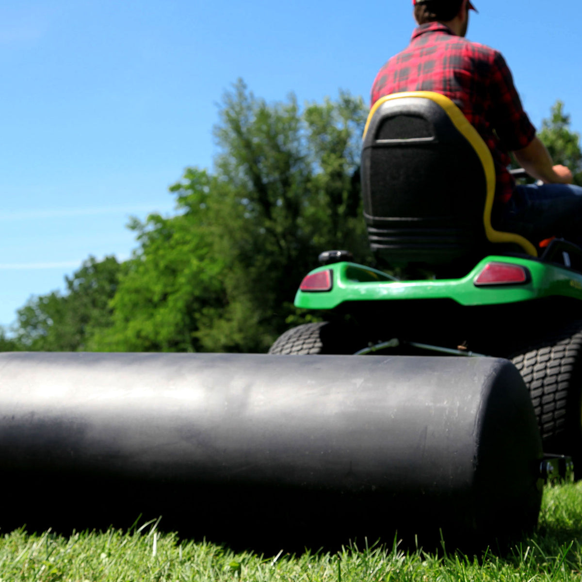 Wearing a red plaid shirt, a person drives a green lawn tractor across the grass, towing the Brinly Parts 54 Gallon Tow-Behind Poly Roller | PRT-482SBH with its rust-free poly drum. Trees and a clear blue sky provide an idyllic backdrop.