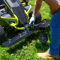 A person in blue jeans and gloves is attaching a Brinly Parts 38 in. DT-38RY Front Mount Dethatcher with a wheel to the front of a green and black RYOBI Electric Riding Mower on grass, focusing on precision lawn care while adjusting the connection with a wrench.