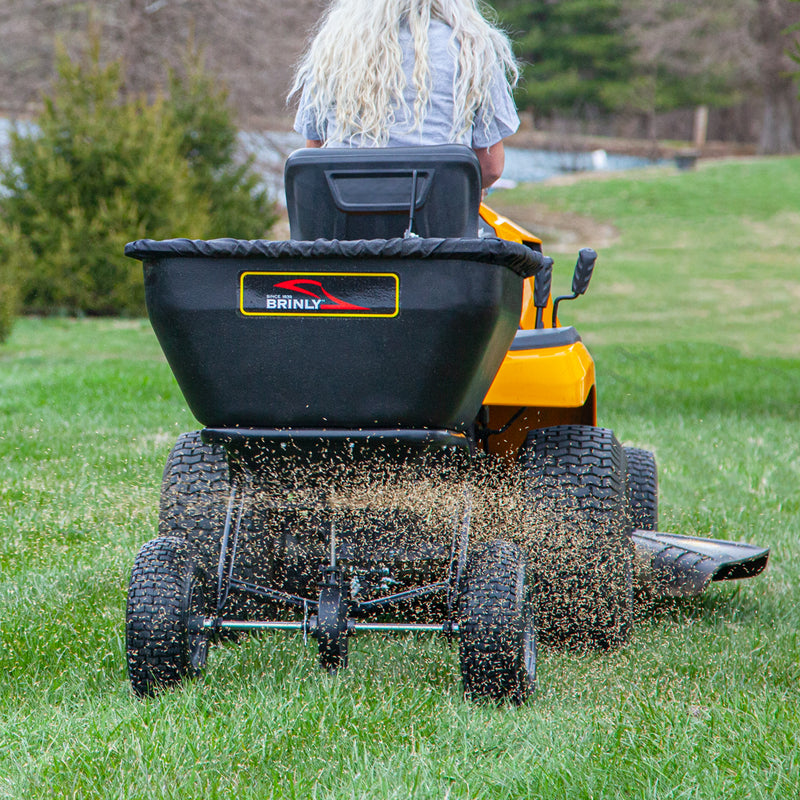 A person with long, curly hair operates a yellow riding mower on the grass, expertly using the Brinly Parts 175 LB. Tow-Behind Deluxe Spreader (BS361BH-A) to efficiently disperse seeds. A black hopper complements the scene against a backdrop of trees and blurred landscape.