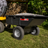 A Brinly Parts 10 Cu. Ft. Poly Cart (PCT-101BH) filled with gravel is attached to a yellow riding lawn mower on grass, with stacked firewood and green trees in the background.