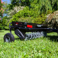 A 40 Combination Aerator Spreader (AS2-40BH-G) by Brinly Parts, featuring pneumatic tires, is placed on vibrant green grass. It boasts a black body with metal spikes and a red handle, surrounded by lush greenery and blooming bushes under a clear blue sky.