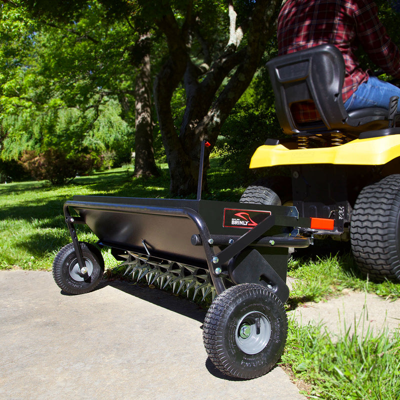 A Brinly Parts 40 Combination Aerator Spreader with Pneumatic Tires (AS2-40BH-G) is hitched to a yellow riding mower on concrete near grass and trees. A person in a plaid shirt operates the mower effortlessly, benefiting from the aerators 100 lb. capacity hopper for efficient soil care.