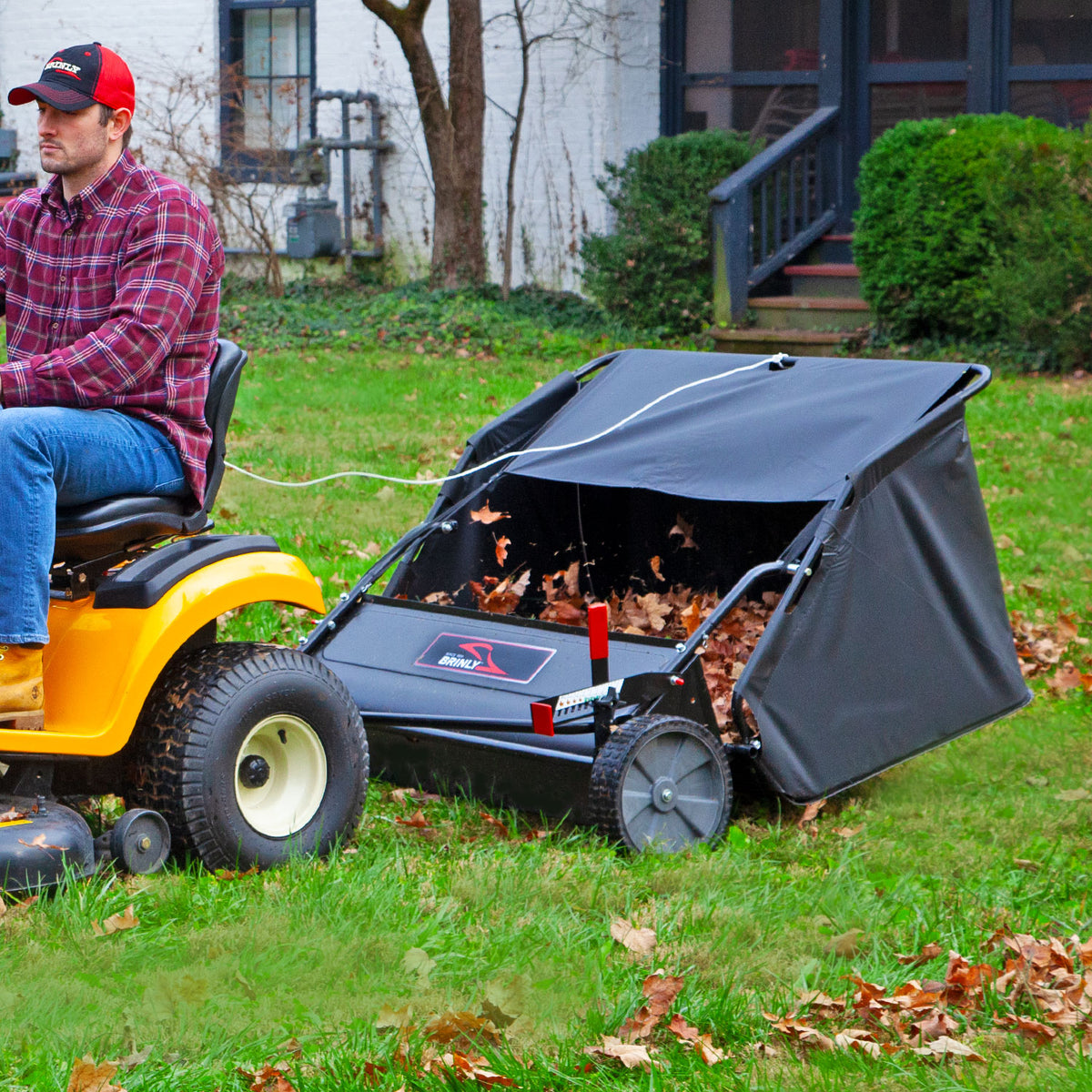 A person wearing a red plaid shirt and cap maneuvers across the grassy yard on a yellow tractor, effortlessly pulling a Brinly Parts 42″ Lawn Sweeper with DOUBLE-HELIX Brushes (Model: LS2-42BH2-G) filled with leaves, while a house and bushes frame the scene in the background.