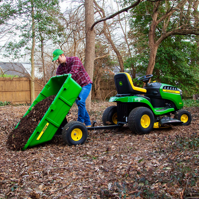 A person in a green cap and red plaid shirt dumps mulch from a John Deere 180 Degree Full Dump Cart, with 650 lb. capacity, attached to a lawn tractor. The outdoor scene is bordered by trees and a wooden fence, with dry leaves covering the ground.