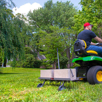 A person in a red cap and black shirt expertly operates a green and yellow lawn tractor with a Brinly Parts 40 Dethatcher in Hammered Gunmetal | DT2-40BH-S. Concrete blocks sit on the cart as large trees and a house adorn the partly cloudy sky backdrop.