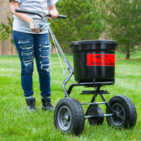 A person in ripped jeans expertly maneuvers a Brinly Parts 50 lb. Push Spreader (P20-500BH) across lush grass. The sturdy spreader, with a black container and red label, has four wheels for perfectly dispensing fertilizer while tall trees offer a serene backdrop to this gardening scene.