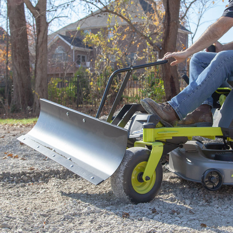 A person skillfully uses a Brinly Parts Front-Mount Blade (FB-42RYZT) on a RYOBI Electric ZTR, efficiently clearing gravel. Trees and houses stand in the background against a clear, bright day.