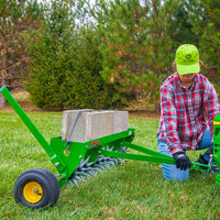 Wearing a green hat and plaid shirt, a person kneels beside the John Deere 40 Spike Aerator with Rigid Steel Weight Tray (model LPSAT40JD/SAT-400JD), set on the grass surrounded by pine trees. Galvanized steel stars are visible, with concrete blocks adding weight on top.