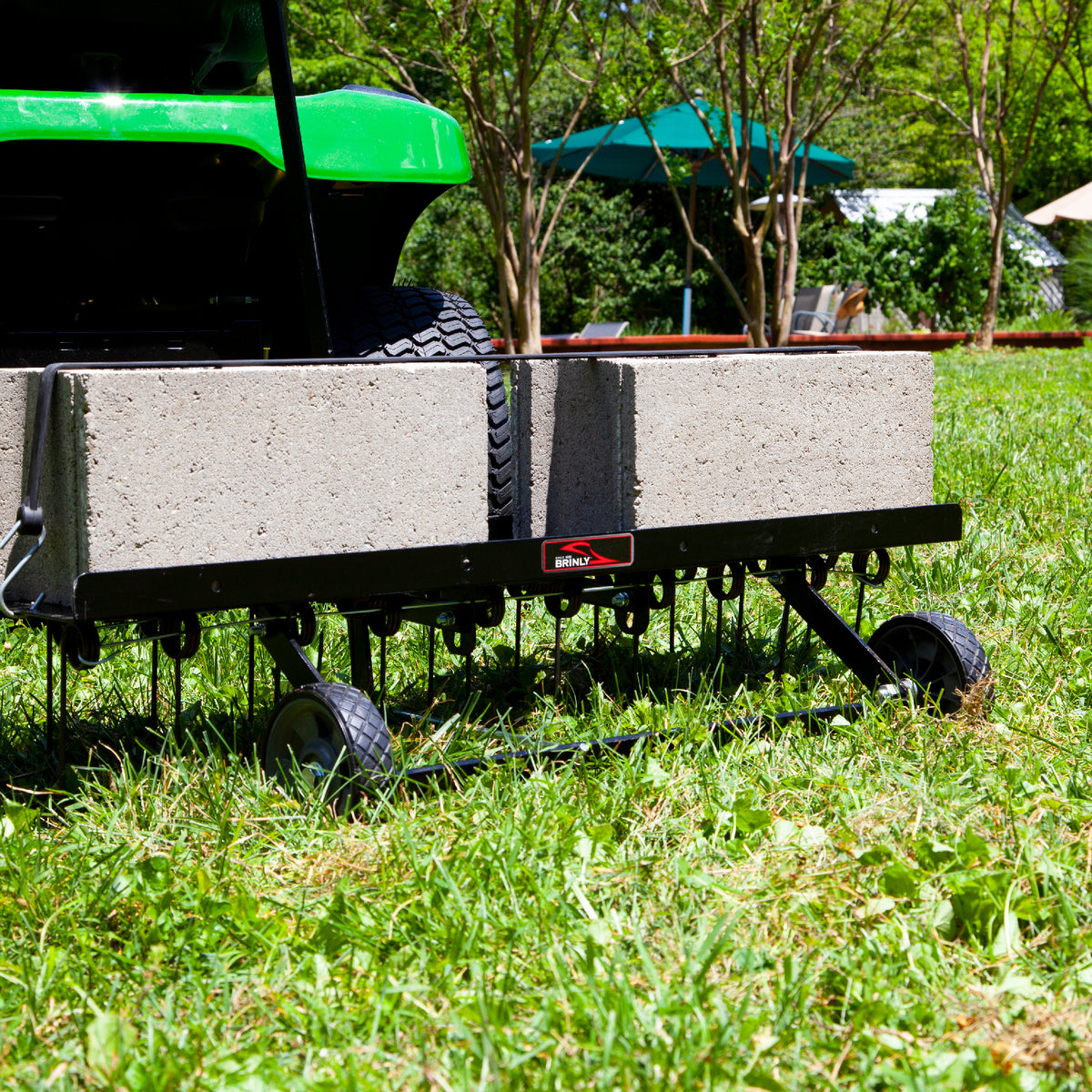 A green vehicle with the Brinly Parts 40″ Tow-Behind Dethatcher (DT2-40BH-G) and an aerator sits on grass, surrounded by trees and a gazebo. Loaded with concrete blocks for weight, its ready for efficient thatch removal.