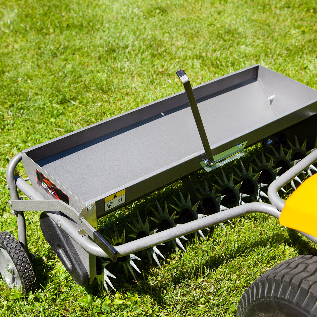 A close-up shows the Brinly Parts 40 Combination Aerator Spreader in Hammered Gunmetal, AS2-40BH-S, featuring black spiked wheels and a rectangular tray on green grass. It attaches to a yellow lawn tractor for efficient soil aeration and lawn maintenance.