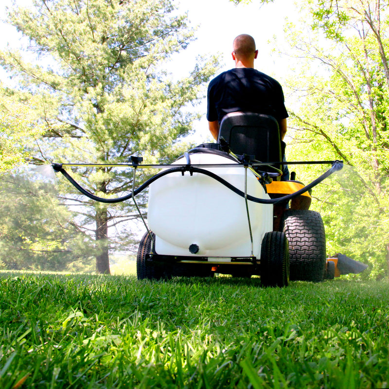 A person in a black shirt uses the Brinly Parts 25 Gallon Tow-Behind Sprayer | ST-251BH with Ultra Lo-Drift™ Spray Tips, efficiently spraying a lush grassy area. The vibrant outdoor scene is bathed in bright sunlight and surrounded by verdant trees.