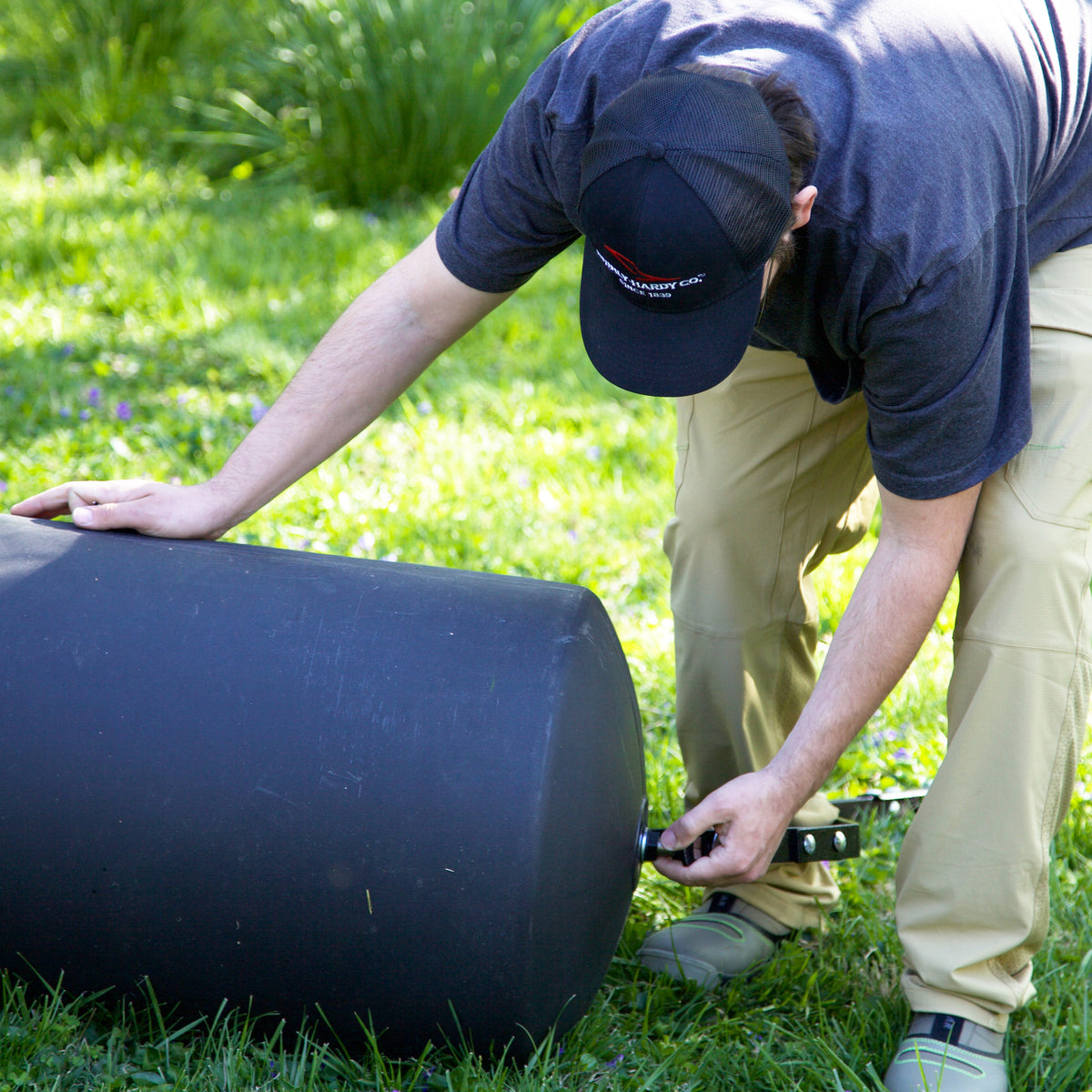 A person wearing a black cap and beige pants kneels on grass, attaching a hose or fitting to a Brinly Parts 28 Gallon Push or Tow Poly Lawn Roller (PRC-243BH).