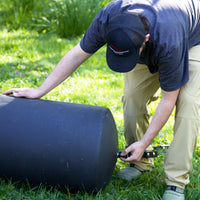 A person wearing a black cap and beige pants kneels on grass, attaching a hose or fitting to a Brinly Parts 28 Gallon Push or Tow Poly Lawn Roller (PRC-243BH).
