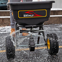 Close-up of a person using the Brinly Parts 70 lbs. Capacity Broadcast Ice Melt Spreader (PS10-70BH) on snow, dispersing salt with its calibration dial and two large wheels. The black hopper features a red and yellow logo as the individual wears brown boots.