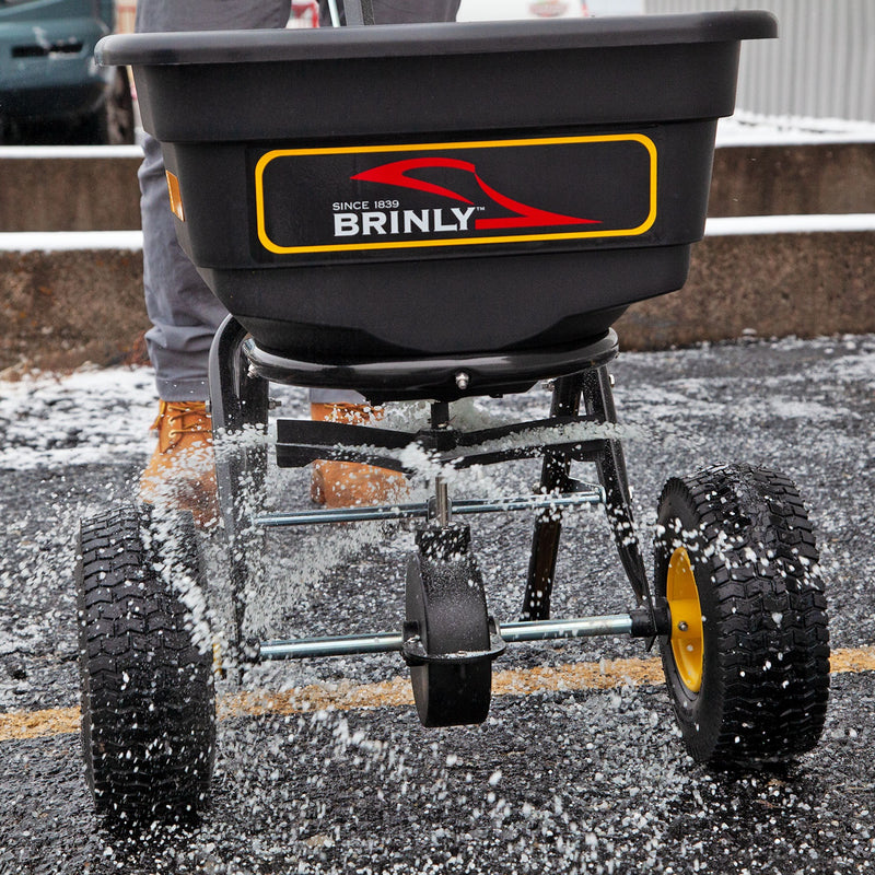 Close-up of a person using the Brinly Parts 70 lbs. Capacity Broadcast Ice Melt Spreader (PS10-70BH) on snow, dispersing salt with its calibration dial and two large wheels. The black hopper features a red and yellow logo as the individual wears brown boots.