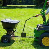 A person operates a green ZTrak lawn tractor with a Brinly Parts 125 LB. Tow-Behind Deluxe Spreader (BS261BH-A) on a grassy field. The rust-proof polyethylene hopper evenly spreads nutrients, while trees create a serene backdrop.