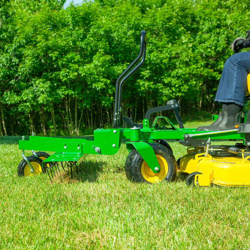 A side view shows an individual using a John Deere Front-Mount 48 ZTR Dethatcher in a grassy field, bordered by dense greenery. Wearing dark pants and boots, their legs are visible as they maneuver effortlessly through the landscape.