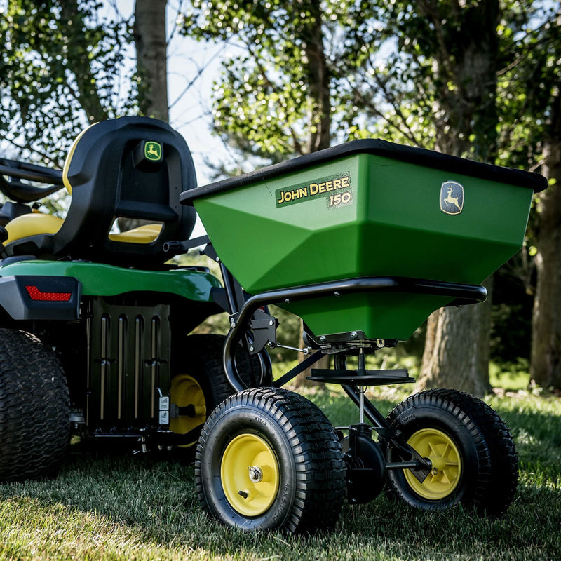 A John Deere 150 lb Tow-Behind Broadcast Spreader with ACCUWAY (LP79931) is connected to a riding lawn mower. Known for PRO-level durability, this green spreader rests on lush grass under a sunny sky, surrounded by trees.