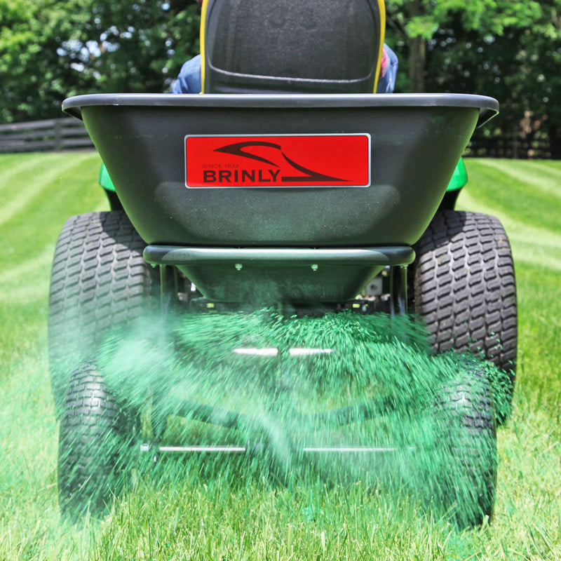 The Brinly Parts 125 lb. Tow-Behind Broadcast Spreader (BS26BH) features a rust-proof polyethylene hopper and large tires, ensuring smooth dispensing of green granules across the lawn when attached to a vehicle, simplifying lawn fertilization amid trees and fences.