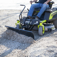 A person uses a Brinly Parts Front-Mount Blade for RYOBI Electric ZTRs (FB-42RYZT) on a green and black Ryobi Electric Zero-Turn Mower to move gravel on dirt. The operator is in jeans, with the mowers seat and controls visible.