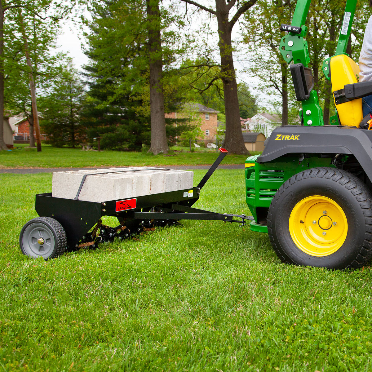 A green lawn tractor with yellow wheels tows a Brinly Parts 48″ Tow-Behind Plug Aerator (PA-482BH) filled with concrete blocks. The steel tines efficiently penetrate the lush grass, enhancing root growth amidst trees and houses.