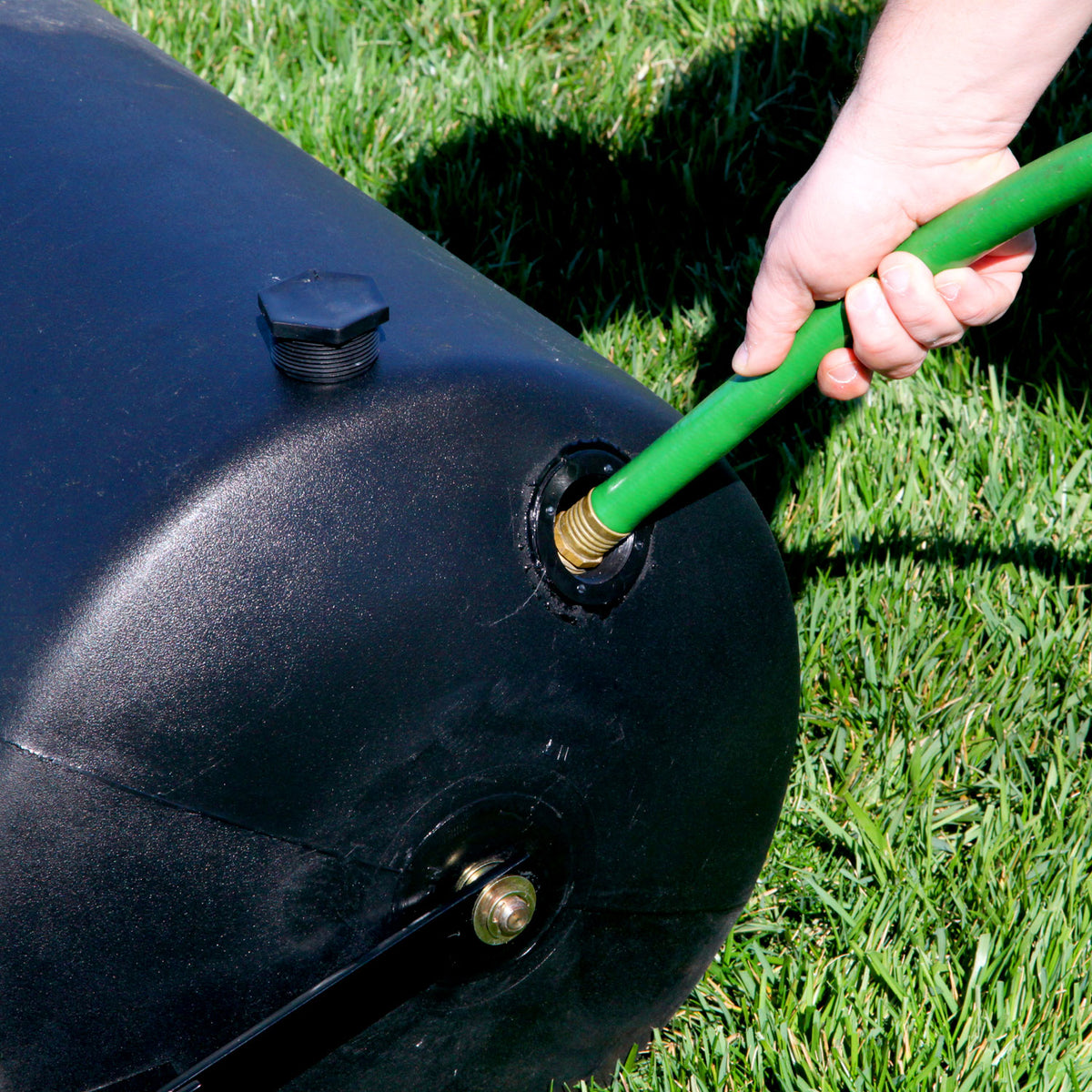 A person inserts a green hose into the Brinly Parts 28 Gallon Push or Tow Poly Lawn Roller on the grass. The cap of the black polyethylene drum is visible near the top, with part of their hand gripping the hose.