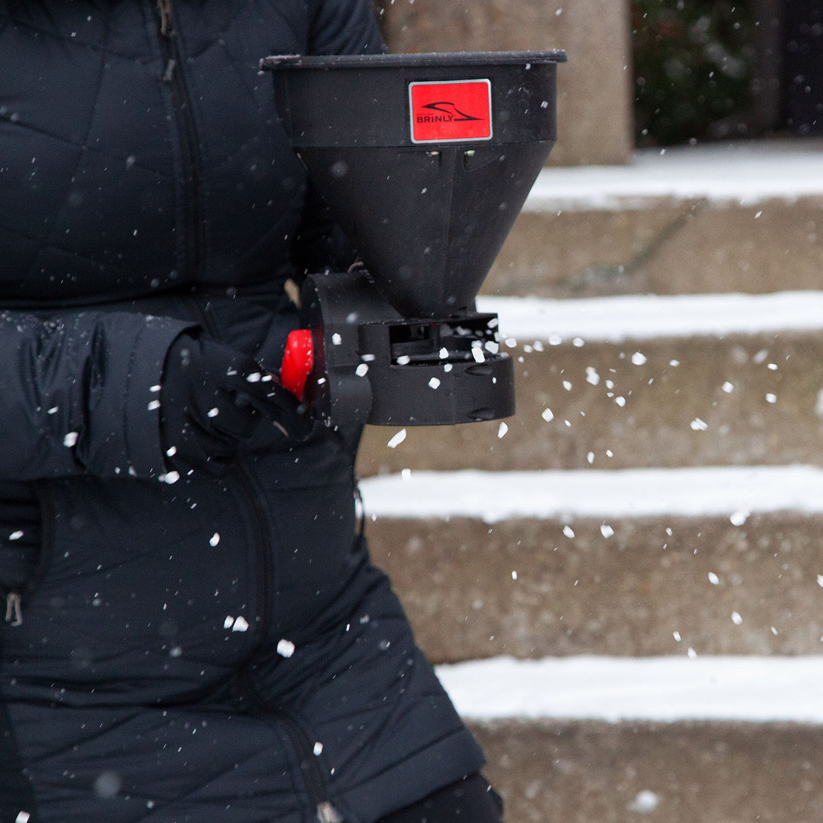 Wearing a dark winter coat, someone uses a Brinly Parts 5lb Capacity Handheld Spreader (HHS3-5BH) to sprinkle ice melt on snow-covered steps. The spreader, black with a red label and knob, glistens as snowflakes dance around.