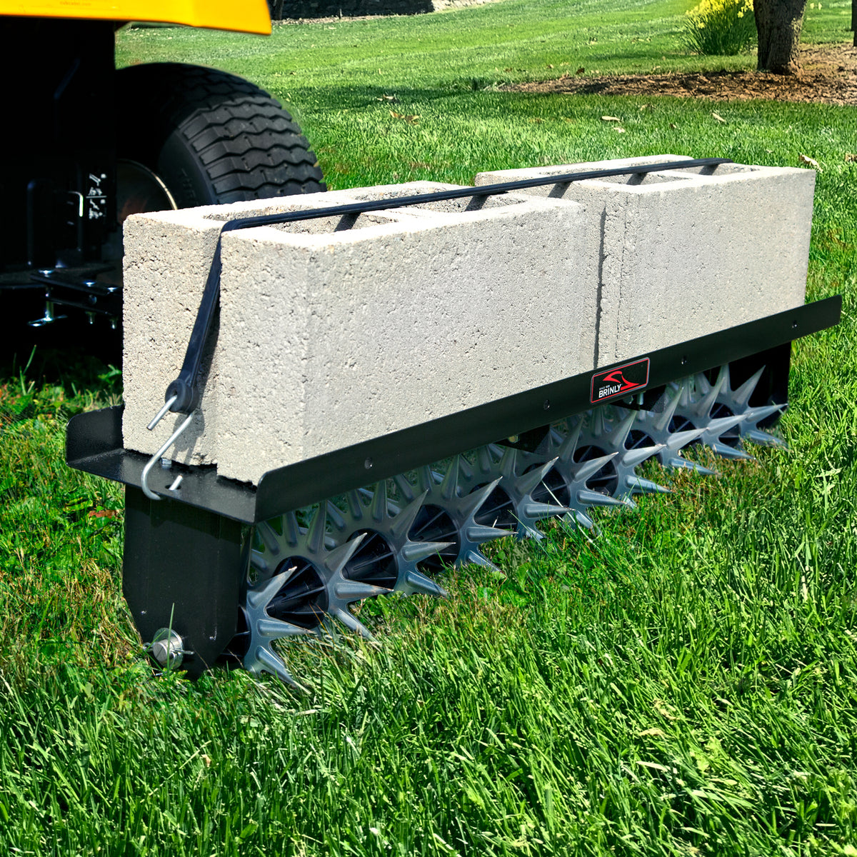 A close-up of the Brinly Parts Spike Aer 40 tow-behind spike aerator with spiked wheels hitched to a vehicle. Two large concrete blocks provide extra weight, improving soil aeration as the spikes penetrate deep into the grass.